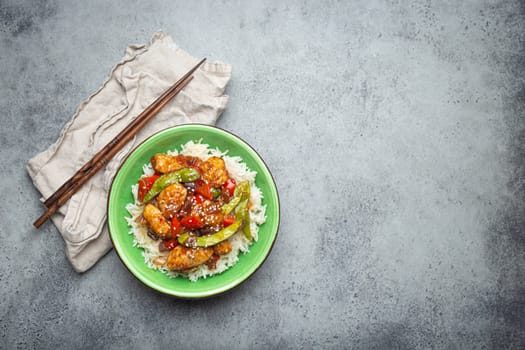 Asian sweet and sour sticky chicken with vegetables stir-fry and rice in ceramic bowl with chopsticks top view on gray rustic stone background, traditional Asian dish. Space for text