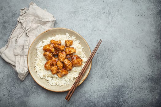 Chinese sweet and sour sticky chicken with sesame seeds and rice on ceramic plate with chopsticks top view on gray rustic stone background, traditional dish of China. Space for text