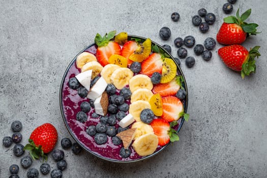 Healthy summer acai smoothie bowl with chia seeds, fresh banana, strawberry, blueberry, cocos, kiwi top view on rustic concrete background with spoon
