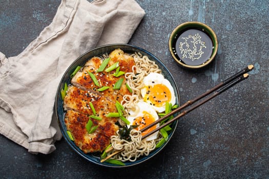 Asian noodles ramen soup with deep fried panko chicken fillet and boiled eggs in ceramic bowl with chop sticks and soy sauce on stone rustic background top view