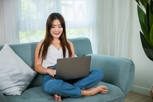 Asian young woman smiling sitting relaxing on sofa using laptop in living room at home, freelance working on computer project, Happy female typing email on notebook computer