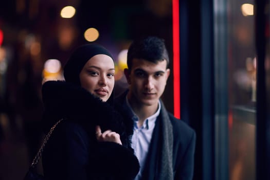 Happy multicultural business couple walking together outdoors in an urban city street at night near a jewelry shopping store window. Successful Arab businessman and European Muslim woman.