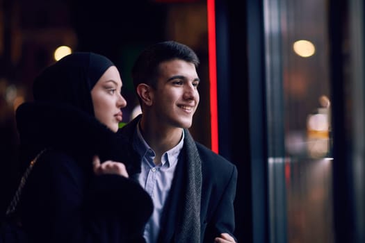 Happy multicultural business couple walking together outdoors in an urban city street at night near a jewelry shopping store window. Successful Arab businessman and European Muslim woman.