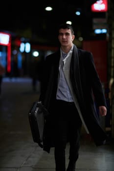 Smiling Meedle Eastern man walking down street near modern office building, freelancer businessman looking away holding mobile phone on busy city street at night