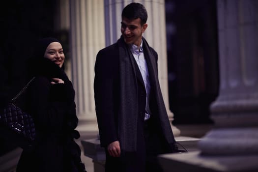 Happy multicultural business couple walking together outdoors in an urban city street at night near a jewelry shopping store window. Successful Arab businessman and European Muslim woman.