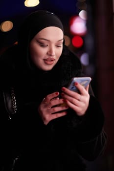 Beautiful and elegant European Muslim Hijabi Business Lady checking her phone and social media on urban city street at night