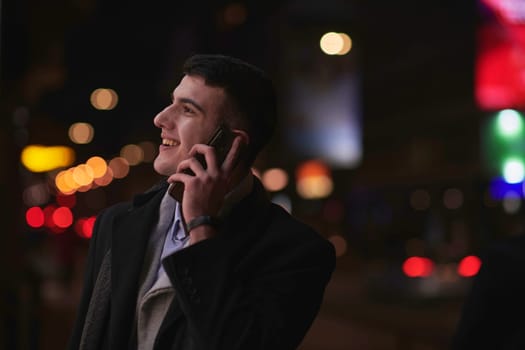 Smiling Meedle Eastern man walking down street near modern office building, freelancer businessman looking away holding mobile phone on busy city street at night