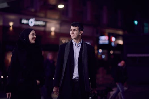 Happy multicultural business couple walking together outdoors in an urban city street at night near a jewelry shopping store window. Successful Arab businessman and European Muslim woman.