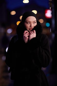 Muslim woman walking on an urban city street on a cold winter night wearing hijab with bokeh city lights in the background