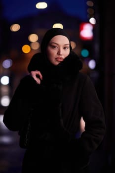 Muslim woman walking on an urban city street on a cold winter night wearing hijab with bokeh city lights in the background