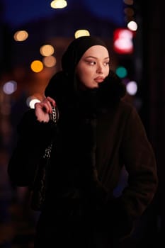 Muslim woman walking on an urban city street on a cold winter night wearing hijab with bokeh city lights in the background