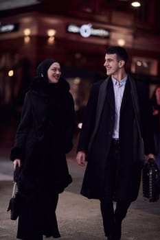 Happy multicultural business couple walking together outdoors in an urban city street at night near a jewelry shopping store window. Successful Arab businessman and European Muslim woman.