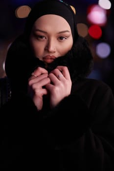 Muslim woman walking on an urban city street on a cold winter night wearing hijab with bokeh city lights in the background