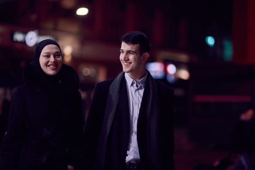 Happy multicultural business couple walking together outdoors in an urban city street at night near a jewelry shopping store window. Successful Arab businessman and European Muslim woman.