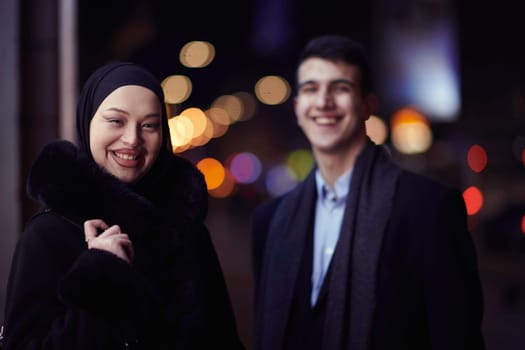 Happy multicultural business couple walking together outdoors in an urban city street at night near a jewelry shopping store window. Successful Arab businessman and European Muslim woman.