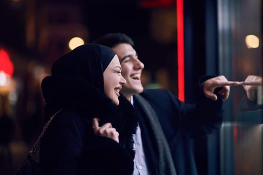 Happy multicultural business couple walking together outdoors in an urban city street at night near a jewelry shopping store window. Successful Arab businessman and European Muslim woman.