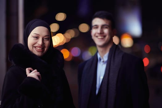 Happy multicultural business couple walking together outdoors in an urban city street at night near a jewelry shopping store window. Successful Arab businessman and European Muslim woman.