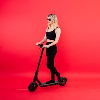 Beautiful emotional young female in a sport clothes on electric scooter on red and white background