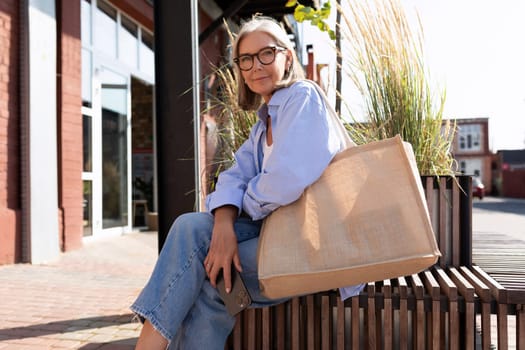 a beautiful mature old woman with gray hair dressed stylishly sits on a bench after shopping.