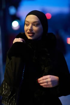 Muslim woman walking on an urban city street on a cold winter night wearing hijab with bokeh city lights in the background