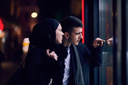 Happy multicultural business couple walking together outdoors in an urban city street at night near a jewelry shopping store window. Successful Arab businessman and European Muslim woman.