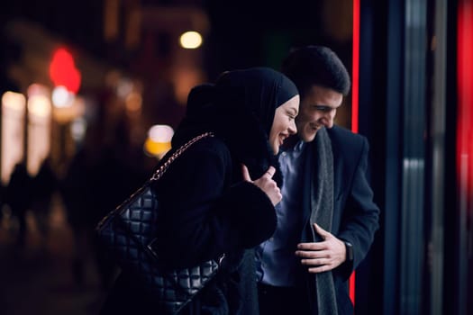 Happy multicultural business couple walking together outdoors in an urban city street at night near a jewelry shopping store window. Successful Arab businessman and European Muslim woman.