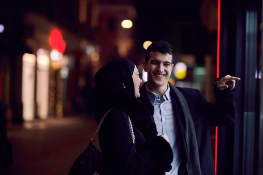 Happy multicultural business couple walking together outdoors in an urban city street at night near a jewelry shopping store window. Successful Arab businessman and European Muslim woman.