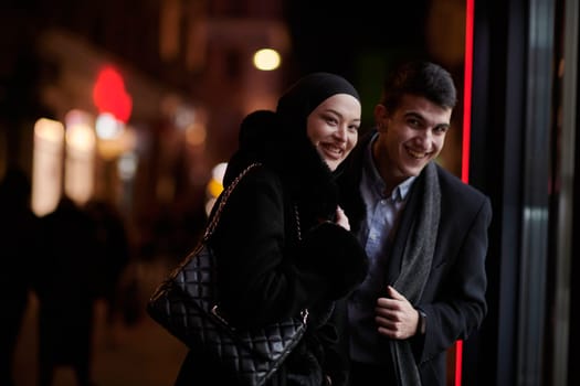 Happy multicultural business couple walking together outdoors in an urban city street at night near a jewelry shopping store window. Successful Arab businessman and European Muslim woman.