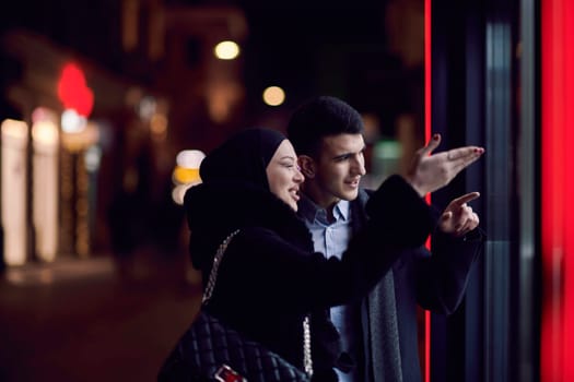 Happy multicultural business couple walking together outdoors in an urban city street at night near a jewelry shopping store window. Successful Arab businessman and European Muslim woman.