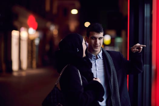 Happy multicultural business couple walking together outdoors in an urban city street at night near a jewelry shopping store window. Successful Arab businessman and European Muslim woman.