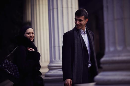 Happy multicultural business couple walking together outdoors in an urban city street at night near a jewelry shopping store window. Successful Arab businessman and European Muslim woman.