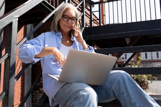 mature grey-haired woman in summer outfit works using a laptop while sitting on the street in the city.