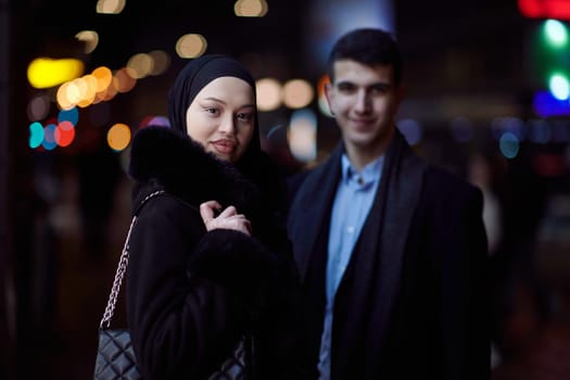 Happy multicultural business couple walking together outdoors in an urban city street at night near a jewelry shopping store window. Successful Arab businessman and European Muslim woman.