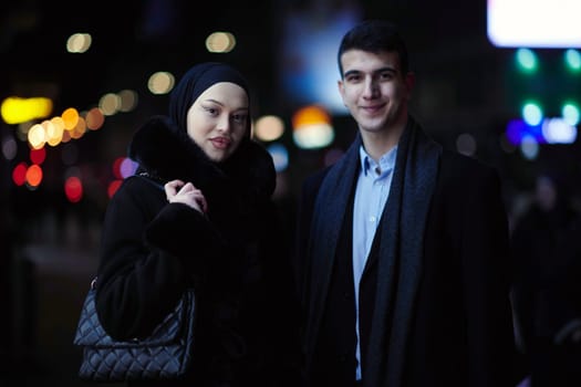 Happy multicultural business couple walking together outdoors in an urban city street at night near a jewelry shopping store window. Successful Arab businessman and European Muslim woman.