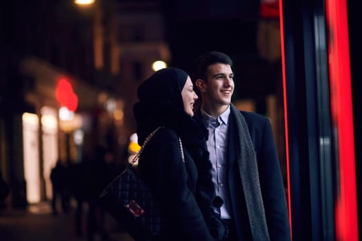 Happy multicultural business couple walking together outdoors in an urban city street at night near a jewelry shopping store window. Successful Arab businessman and European Muslim woman.