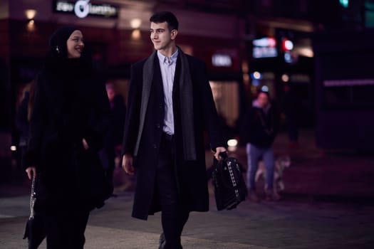 Happy multicultural business couple walking together outdoors in an urban city street at night near a jewelry shopping store window. Successful Arab businessman and European Muslim woman.