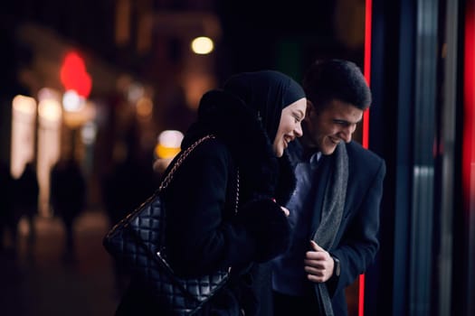 Happy multicultural business couple walking together outdoors in an urban city street at night near a jewelry shopping store window. Successful Arab businessman and European Muslim woman.