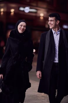 Happy multicultural business couple walking together outdoors in an urban city street at night near a jewelry shopping store window. Successful Arab businessman and European Muslim woman.