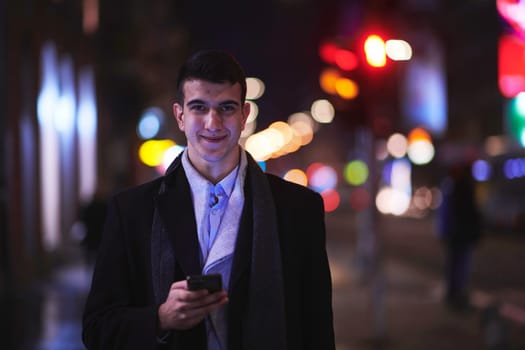 Smiling Meedle Eastern man walking down street near modern office building, freelancer businessman looking away holding mobile phone on busy city street at night