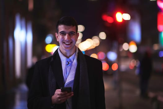 Smiling Meedle Eastern man walking down street near modern office building, freelancer businessman looking away holding mobile phone on busy city street at night