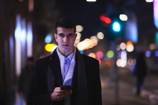 Smiling Meedle Eastern man walking down street near modern office building, freelancer businessman looking away holding mobile phone on busy city street at night