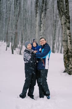 Smiling parents with a small child in their arms stand under snowfall in the forest. High quality photo