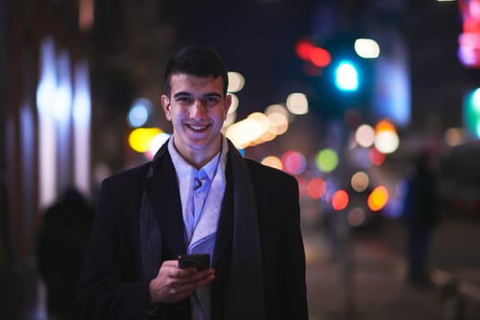 Smiling Meedle Eastern man walking down street near modern office building, freelancer businessman looking away holding mobile phone on busy city street at night