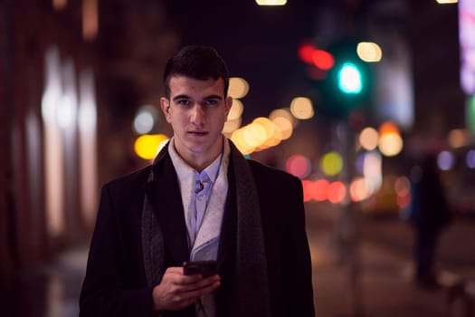 Smiling Meedle Eastern man walking down street near modern office building, freelancer businessman looking away holding mobile phone on busy city street at night