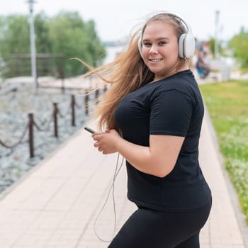 Young fat woman in headphones walks outdoors and listens to music on a smartphone.