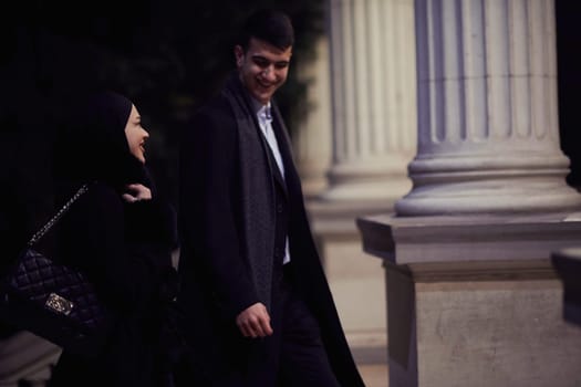 Happy multicultural business couple walking together outdoors in an urban city street at night near a jewelry shopping store window. Successful Arab businessman and European Muslim woman.