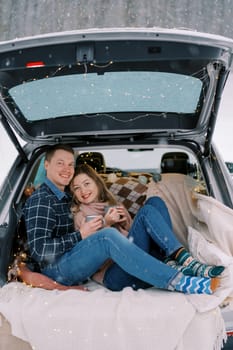 Smiling couple leaning against each other while sitting with mugs of coffee on a blanket in the trunk of a car. High quality photo