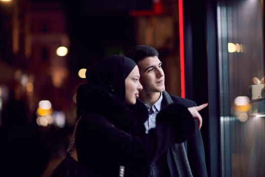 Happy multicultural business couple walking together outdoors in an urban city street at night near a jewelry shopping store window. Successful Arab businessman and European Muslim woman.