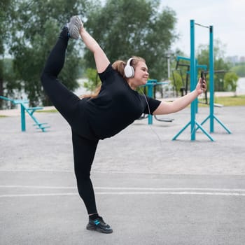 Beautiful fat young woman in headphones makes a figure skater and takes a selfie. A chubby smiling girl listens to music and warms up outdoors. Sports for weight loss