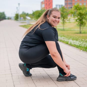 A fat woman in a tracksuit crouches down and ties her shoelaces outdoors